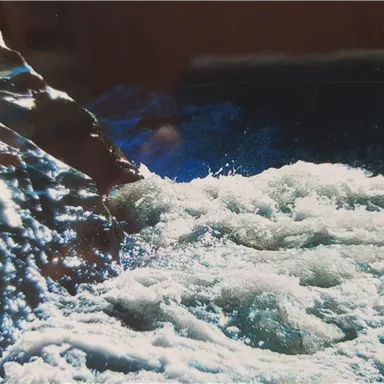 Ein rauschender Wasserfluss trifft auf einen felsigen Uferbereich. Das Wasser schimmert in verschiedenen Blau- und Grüntönen.
