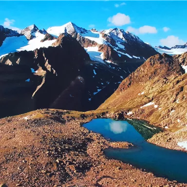 Eine beeindruckende Berglandschaft mit schneebedeckten Gipfeln und einem klaren Blau eines kleinen Sees. Der Himmel ist strahlend blau mit wenigen Wolken.