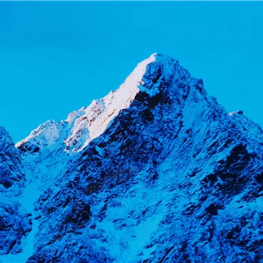 Ein schneebedeckter Berggipfel unter einem klaren blauen Himmel. Die Spitze ist von Sonnenlicht beleuchtet.