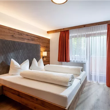 A cozy bedroom with wooden details and two single beds. Large windows let in plenty of light, and the curtains are in a warm tone.