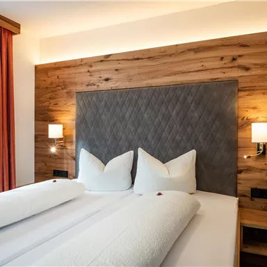 A modern bedroom with a double bed and wooden paneling. The walls are light and illuminated with elegant lamps.