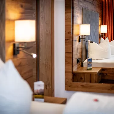 A comfortable hotel room with wooden details and a large mirror. In the background, a bed and cozy cushions are visible.