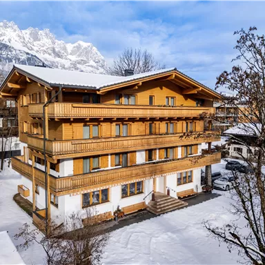 A cozy wooden building in the snow, surrounded by treetops. In the background, majestic mountains are visible.