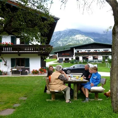 A group of people is sitting at a table outdoors, enjoying a meal. In the background, there is a traditional house and a beautiful mountain landscape.