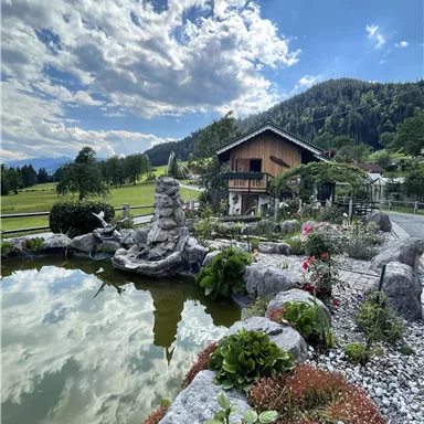 An idyllic garden with a pond and a stone sculpture. In the background, there is a traditional wooden house, surrounded by green meadows and mountains.