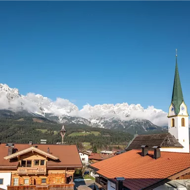 Eine malerische Landschaft mit traditionellen Gebäuden und einer auffälligen Kirche. Im Hintergrund sind Berge mit schneebedeckten Gipfeln und blauem Himmel zu sehen.