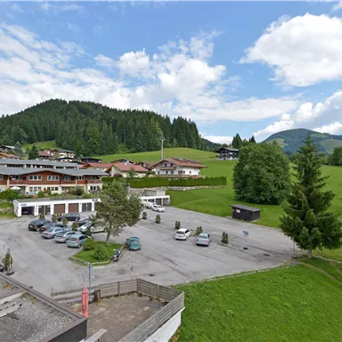 Eine ruhige Landschaft mit grünen Wiesen und Bergen im Hintergrund. Mehrere Häuser und ein Parkplatz sind sichtbar.