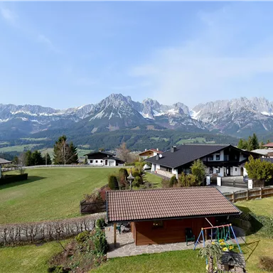 A picturesque landscape with snow-covered mountains in the background. In the foreground, meadows and traditional houses can be seen.