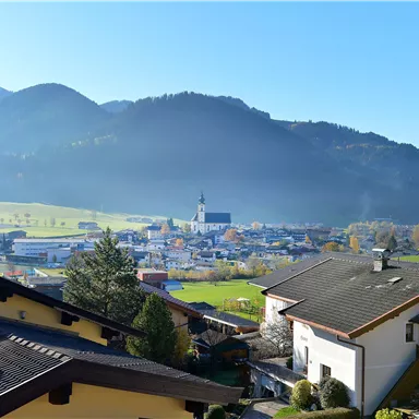 A picturesque view of a village surrounded by mountains. The sky is clear and blue, and the houses are arranged in a peaceful landscape.