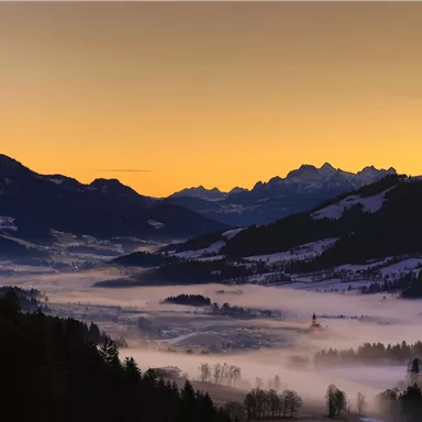 A picturesque mountain landscape with gentle hills and snow-capped peaks. The sky glows in warm hues during dawn.