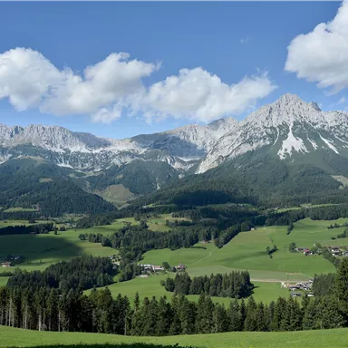 Eine beeindruckende Berglandschaft mit hohen Gipfeln und grünen Wiesen. Strahlend blauer Himmel mit wenigen Wolken vervollständigt das Bild.