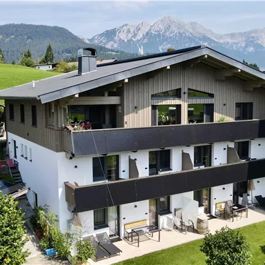 Ein modernes Haus mit mehreren Balkonen, die mit bunten Blumen dekoriert sind. Vor dem Gebäude befinden sich Sitzgelegenheiten und Liegen im Garten.