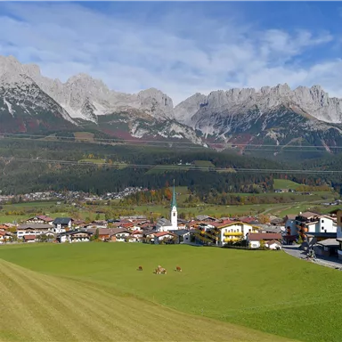 Eine malerische Berglandschaft mit majestätischen Gipfeln und einer kleinen Ortschaft im Vordergrund. Die Wiesen sind grün und es herrscht ein klarer blauer Himmel.