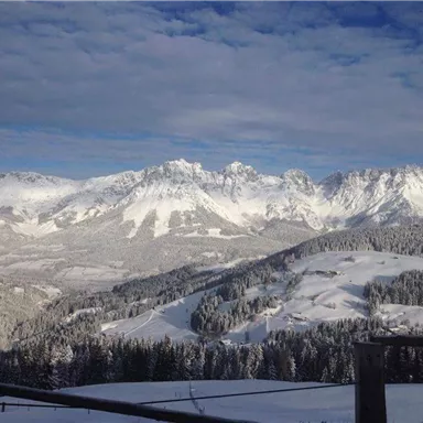 Eine winterliche Berglandschaft mit schneebedeckten Gipfeln und Wäldern. Der Himmel ist teilweise bewölkt und verleiht der Szene eine ruhige Atmosphäre.