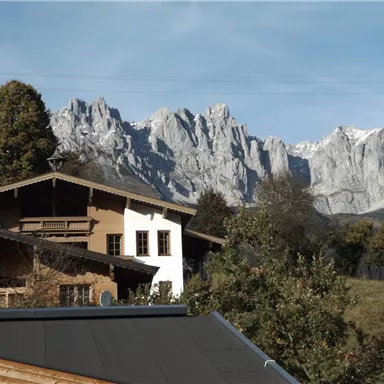 Eine malerische Berglandschaft mit schneebedeckten Gipfeln im Hintergrund. Im Vordergrund sind traditionelle Häuser und grüne Bäume zu sehen.