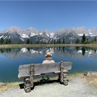 Eine Person sitzt auf einer Bank am Ufer eines klaren Sees. Im Hintergrund sind majestätische Berge und ein blauer Himmel zu sehen.
