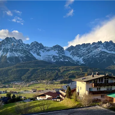 Eine malerische Berglandschaft mit schneebedeckten Gipfeln unter einem klaren blauen Himmel. Im Vordergrund sind charmante Gebäude und grüne Wiesen zu sehen.