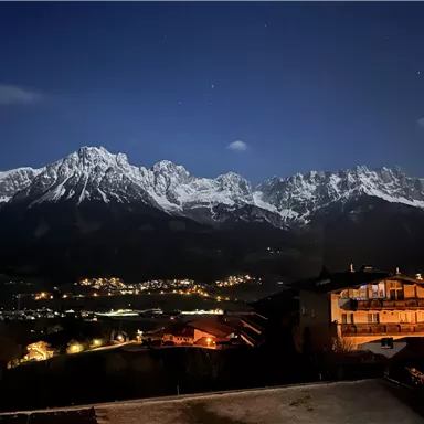 Eine beeindruckende Berglandschaft bei Nacht mit schneebedeckten Gipfeln. Im Vordergrund sind beleuchtete Häuser zu sehen.