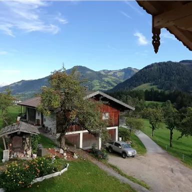 Ein idyllisches Bauernhaus inmitten von grünen Wiesen und Bergen. Der Himmel ist blau und klar, was eine friedliche Atmosphäre schafft.