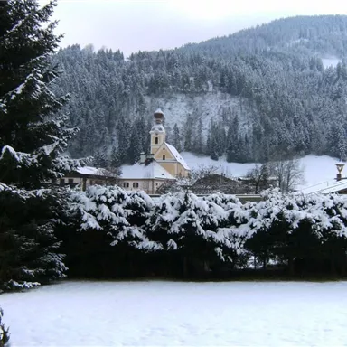 A snowy landscape with pine trees and a small village. In the background, a church is visible, surrounded by snow-covered mountains.