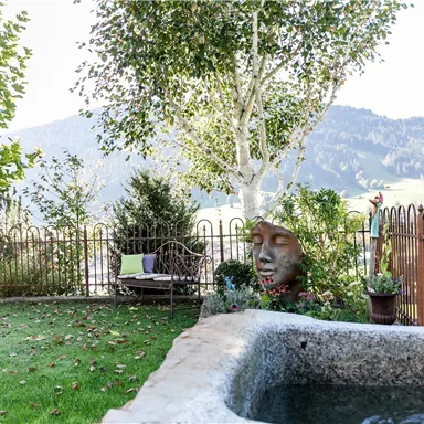 Ein schöner Garten mit einer Steinfigur und einem kleinen Wasserbecken. Im Hintergrund sind Berge und eine freundliche Umzäunung zu sehen.