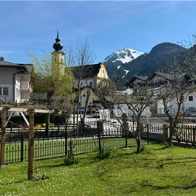 An idyllic village with beautiful houses and a church tower. In the background, snow-covered mountains and a clear blue sky can be seen.