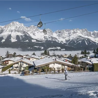 Eine schneebedeckte Landschaft mit einem Skifahrer und malerischen Berggipfeln im Hintergrund. Die Häuser sind im Vordergrund sichtbar, während die Seilbahnen über die Szene fahren.