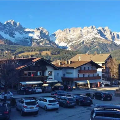 Ein malerisches Dorf mit traditionellen Häusern und einem Parkplatz. Im Hintergrund erheben sich majestätische Berge mit schneebedeckten Gipfeln.