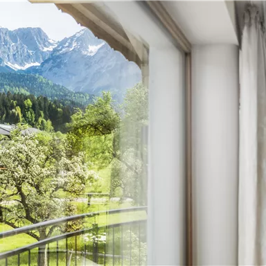 Ein Blick aus einem Fenster mit einer schönen Berglandschaft im Hintergrund. Im Vordergrund sieht man Bäume und eine gepflegte Wiese.