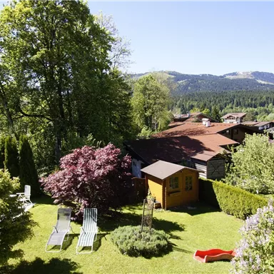 Ein grüner Garten mit bunten Blumen und einem kleinen Holzhaus. Im Hintergrund sind Hügel und eine klare blauen Himmel zu sehen.