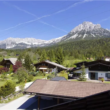 Eine malerische Landschaft mit Bergblick und traditionellen Häusern. Grüne Wiesen und ein blauer Himmel verleihen der Szene eine friedliche Atmosphäre.