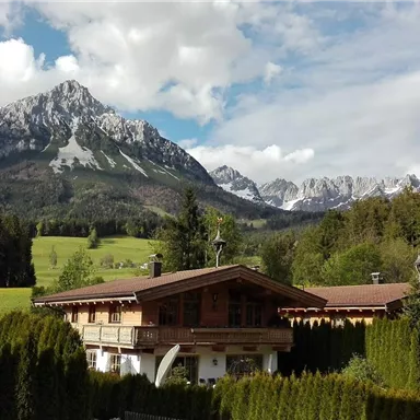 Eine idyllische Berglandschaft mit schneebedeckten Gipfeln und sattgrünen Wiesen. Im Vordergrund steht ein gemütliches Holzhaus umgeben von Bäumen.