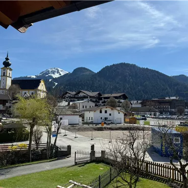 A picturesque landscape with houses, trees, and mountains in the background. The sky is clear and blue, and the scene radiates tranquility and idyll.