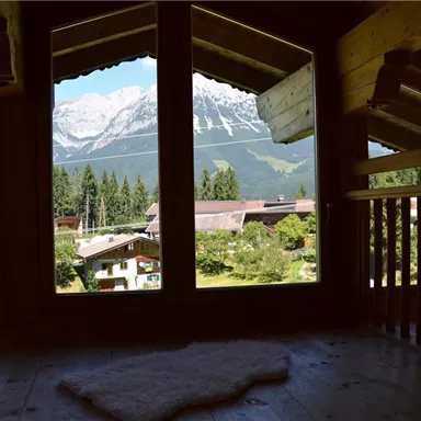 A view from a wooden house onto snow-covered mountains and green meadows. In the foreground, window frames and a carpet are visible.