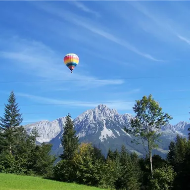 Ein buntes Heißluftballon schwebt über einer malerischen Berglandschaft. Grünes Gras und hohe Bäume umrahmen die Szene.