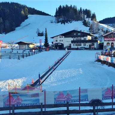 Eine verschneite Landschaft mit einem präparierten Skikurs. Im Hintergrund sind traditionelle alpine Gebäude und Bäume zu sehen.