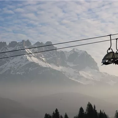 Eine Seilbahn schwebt über einem gebirgigen Landschaftsbild. Im Hintergrund sind schneebedeckte Gipfel und eine bewölkte Himmel sichtbar.