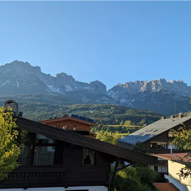 A picturesque mountain landscape with high peaks and a clear blue sky. In the foreground, traditional houses and trees can be seen.