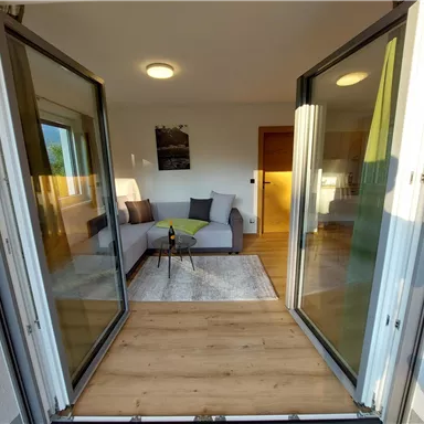 A bright living space with a cozy couch and a table. The doors open to an outdoor area with a wooden floor.