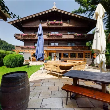 A traditional wooden house with blooming balcony boxes, surrounded by a beautiful green landscape. In the foreground are wooden furniture and a large sunshade.
