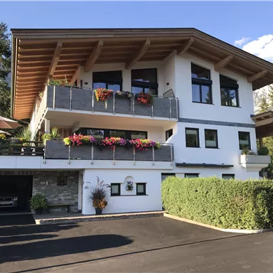 Ein modernes Haus mit einem großen Balkon und bunten Blumen. Umgeben von Bäumen und einem klaren Himmel.