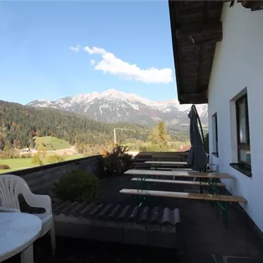 Eine Terrasse mit Sitzmöglichkeiten und Blick auf die Berge. Die Umgebung ist grün und sonnig, mit schneebedeckten Gipfeln im Hintergrund.