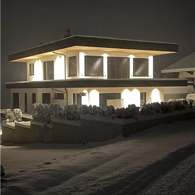 A modern house, illuminated at night, stands in a snowy landscape. The snow reflects the light and creates a tranquil atmosphere.