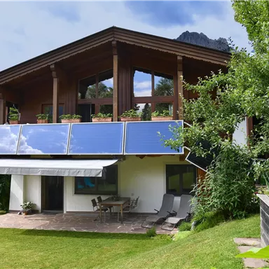 Ein modernes Haus mit Solarpanelen auf dem Dach, umgeben von einem grünen Garten. Der Himmel ist teilweise bewölkt und es gibt einen gemütlichen Außenbereich mit Sitzgelegenheiten.