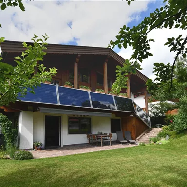 Ein modernes Haus mit Holzverkleidung und Solarpanelen auf dem Dach. Der Garten ist grün und einladend, mit Sitzmöglichkeiten im Freien.