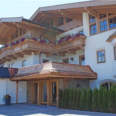 Ein charmantes Gebäude im alpinen Stil mit Holzverkleidung und blühenden Balkonkästen. Die Fassade ist hell und freundlich, umgeben von gepflegtem Grün.