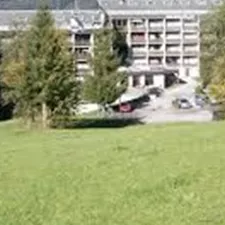Ein großes Gebäude steht auf einer Wiese, umgeben von Bäumen und Hügeln. Im Hintergrund sind grüne Berge sichtbar.
