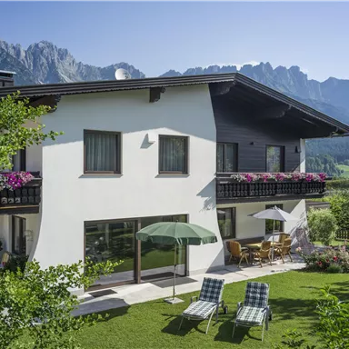 A modern house with a well-kept garden and a view of the mountains. On the terrace, there are deck chairs and a sunshade.