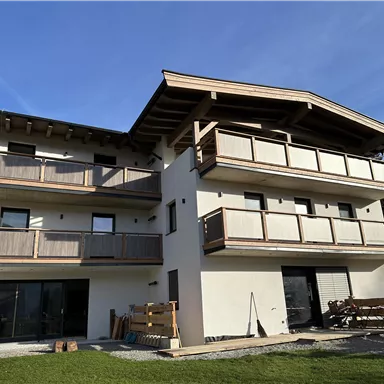 A modern building with several balconies and a wooden roof. It is situated in a green environment with trees in the background.