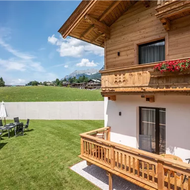 A cozy wooden house with a balcony and flowers. The garden is green and furnished with terrace furniture to enjoy the view.